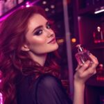 Mulher de cabelos ruivos segurando um perfume, com prateleiras cheias de produtos ao fundo.